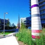 A Set Of Spire Sculptures Installed In The Ground Along A Path In Between Two Buildings Are Tapered From A Wide Base To The Pointed Tip And Are Wrapped With Horizontal Rainbow Coloured Bands