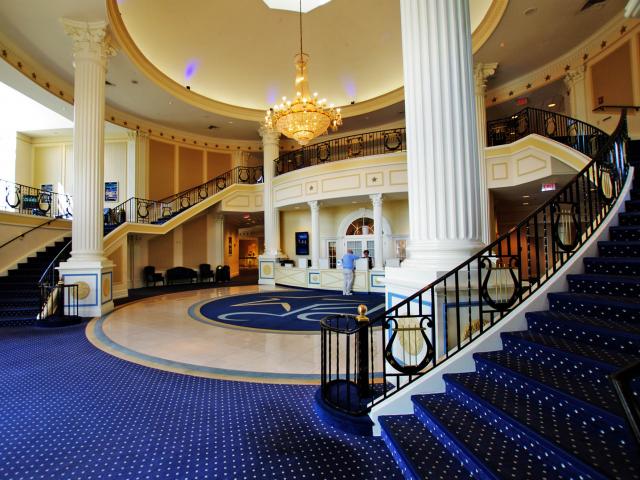 A Grand Lobby With 2 Staircases Wrapping Around Each Side Ascending 2 Floors To Meet At A Large Central Landing Above A Service Desk