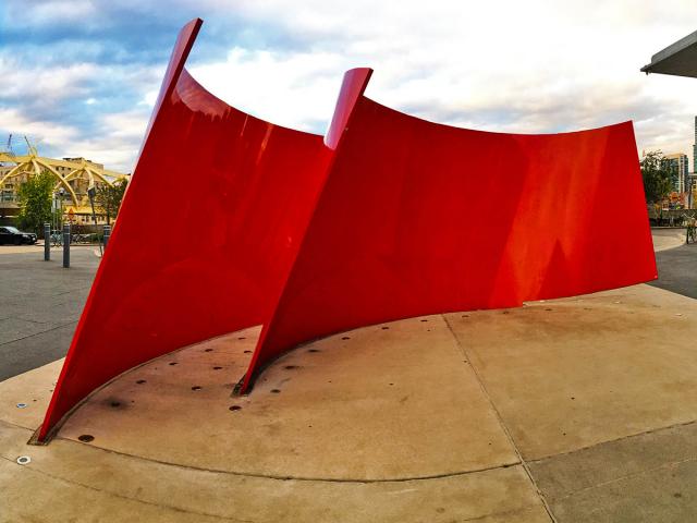 A Bright Shiny Red Metal Sculpture Resembling Two Pieces Of Ribbon In A Semi-Circle Sits In Front Of A Building Entrance