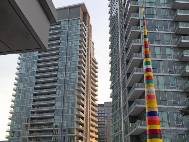Looking Up At A Spire Sculpture Which Is Tapered From A Wide Base To The Pointed Tip And Wrapped With Horizontal Rainbow-Coloured Bands In Front Of A Building